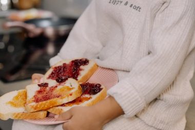 A child in a cozy sweater holds a plate of jam sandwiches indoors.